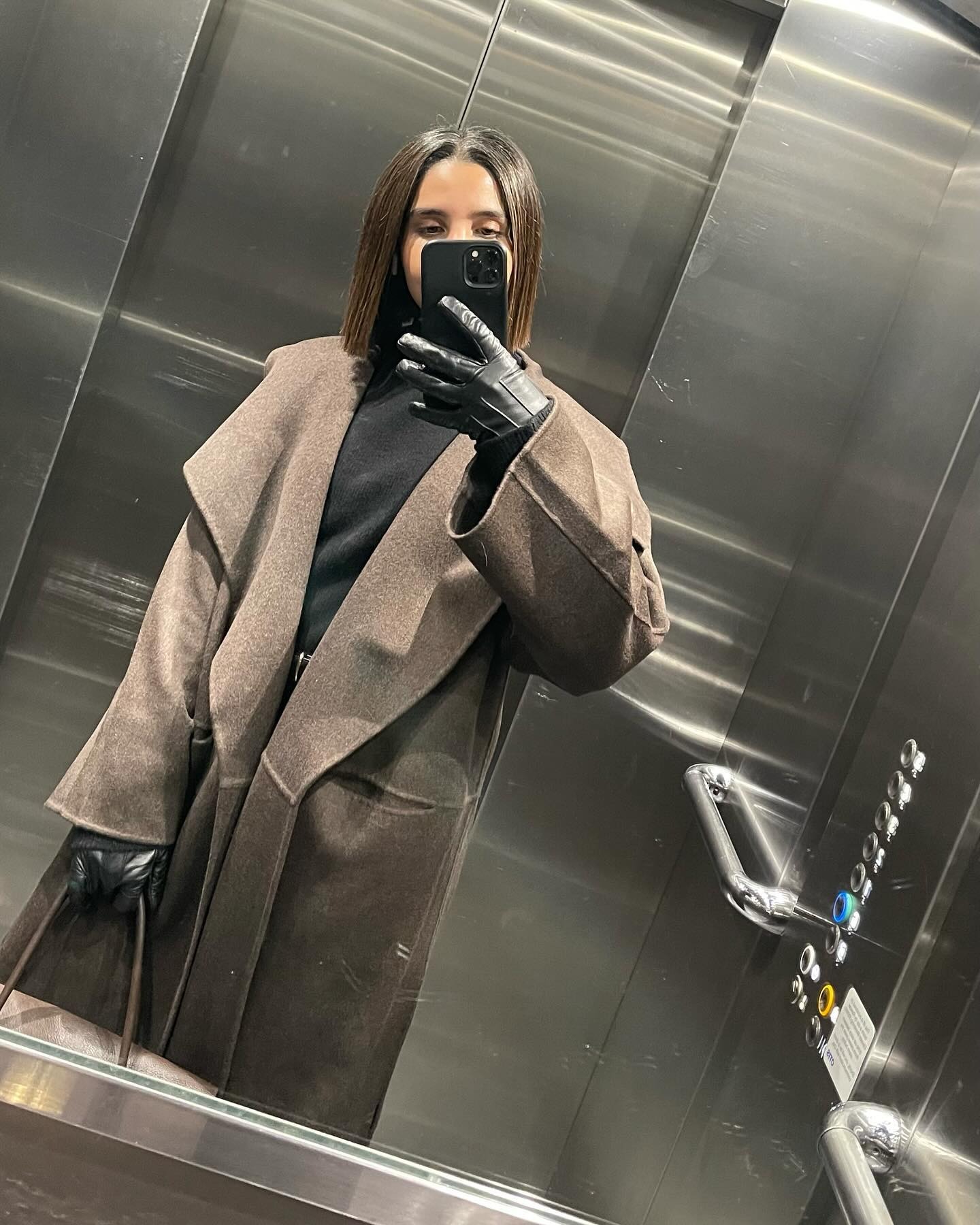 Humaa Hussain, koyu kahverengi bob saç kesimi ve kahverengi bir paltoyla asansörde selfie çekiyor