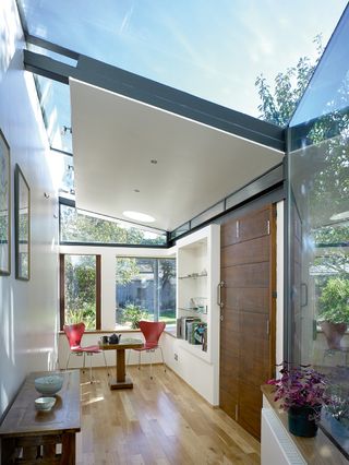 a modern single-storey side extension with glass roof and walls, wooden floor, a red dining set at the far end and a large wooden front door