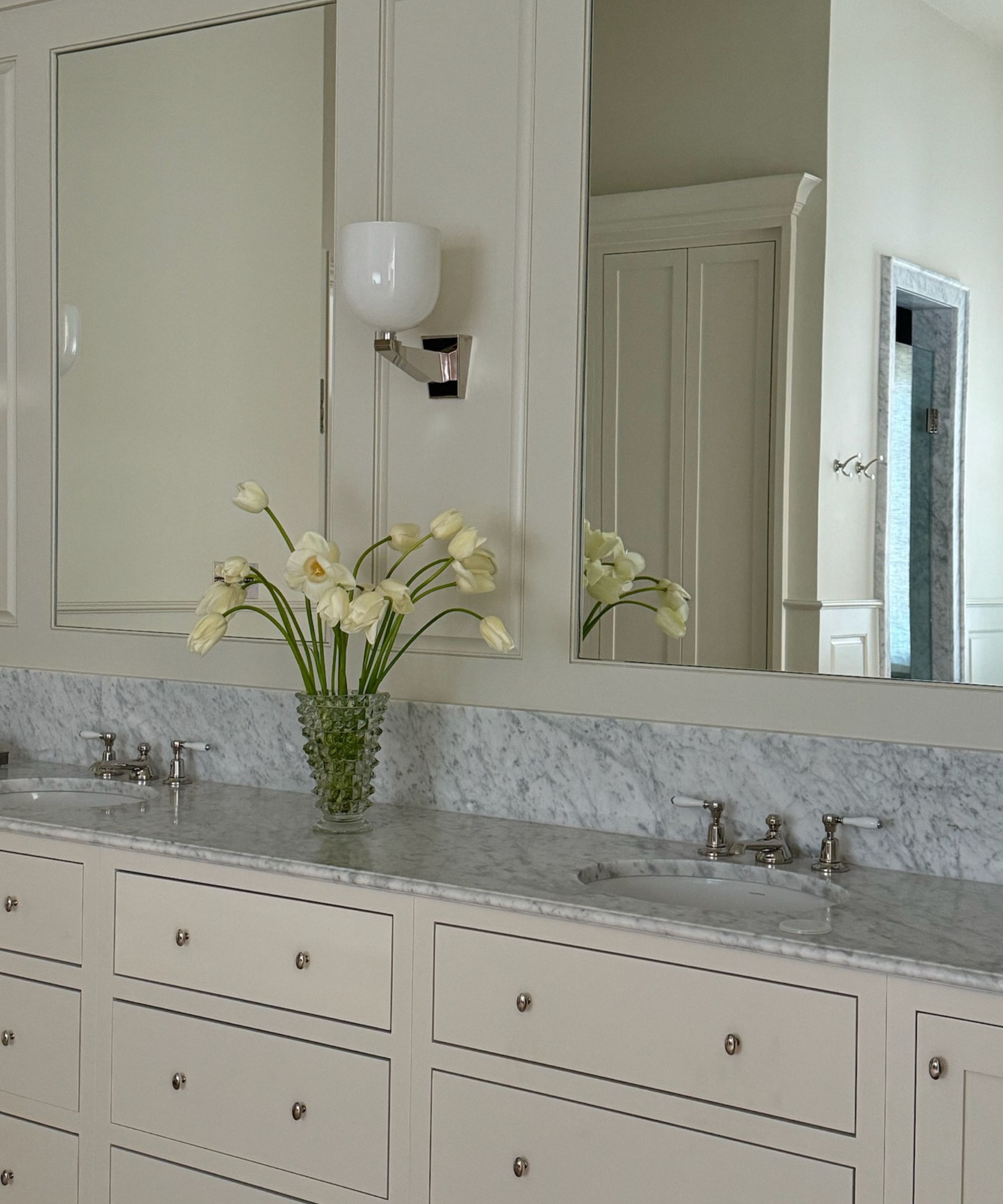 Cream bathroom with marble counterop and backsplash. Silver and white glass wall lamp beside mirrored cream cabinetry. Yellow flowers in a glass vase on counter