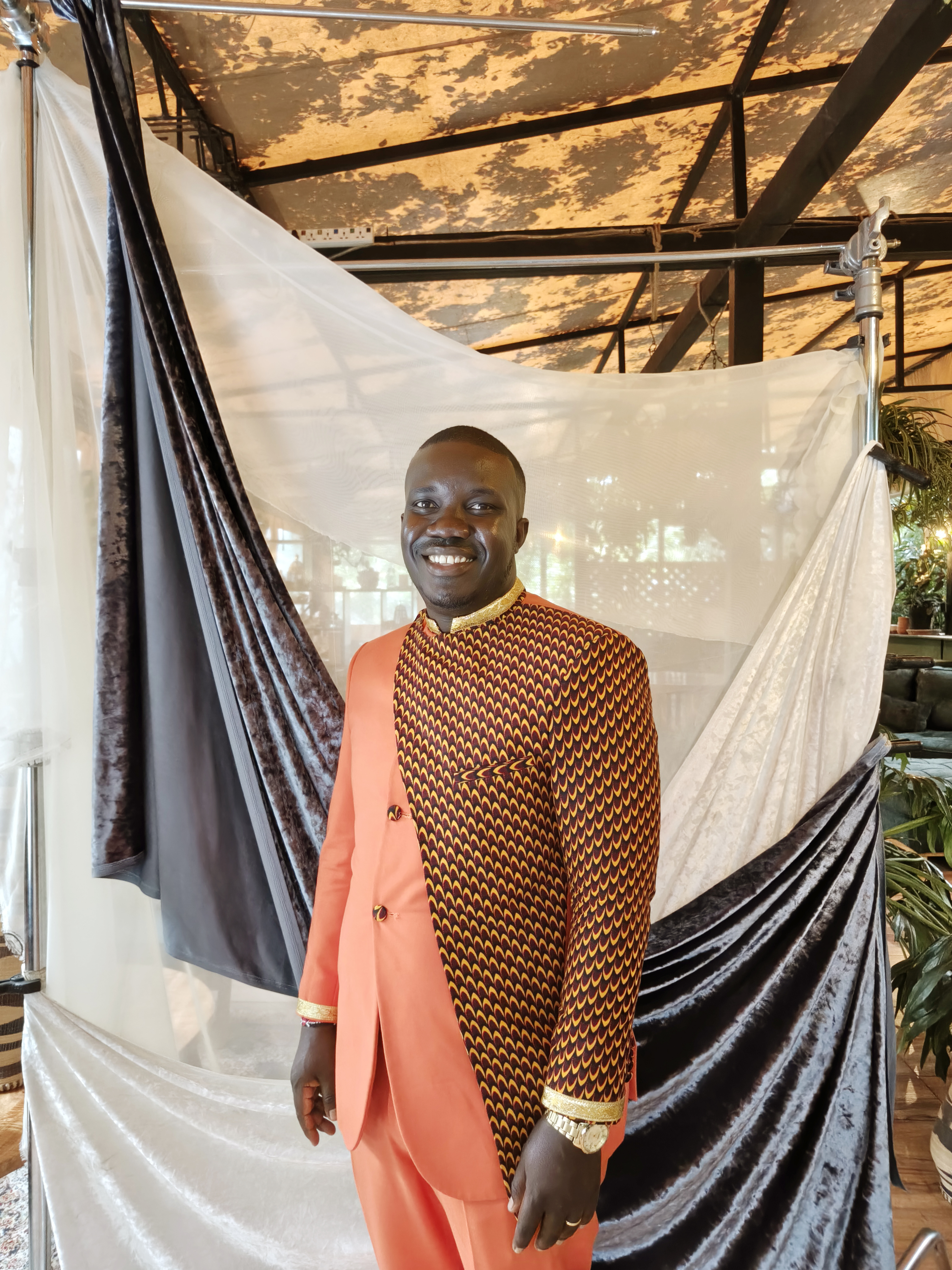 A smiling man in a coral suit with a patterned front panel and gold trim poses in front of draped white and dark velvet fabrics inside a greenhouse-style venue.