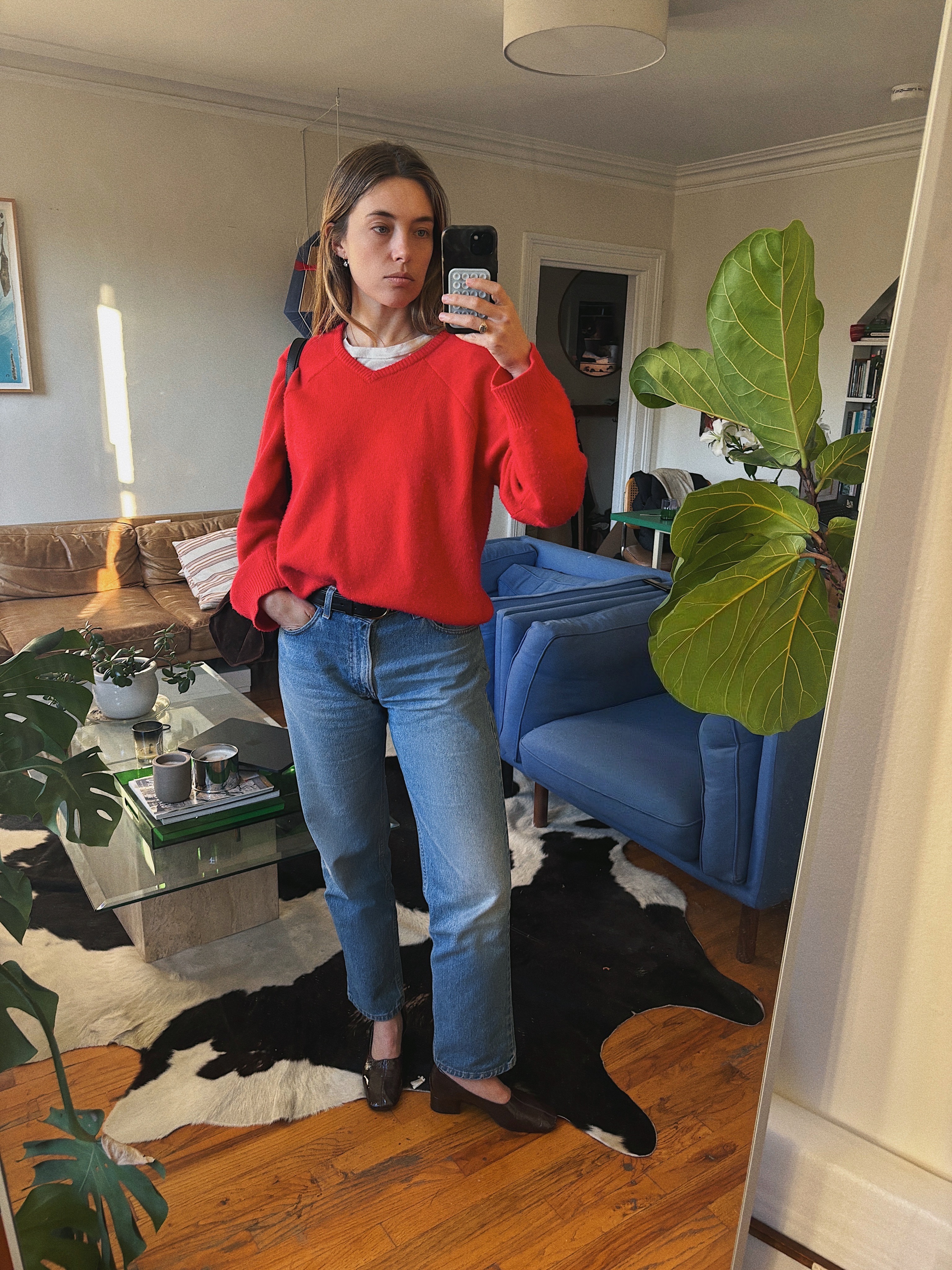 Ameilia Madden in a red v neck sweater, white tee shirt, baggy blue denim jeans, brown flat shoes holding an iPhone
