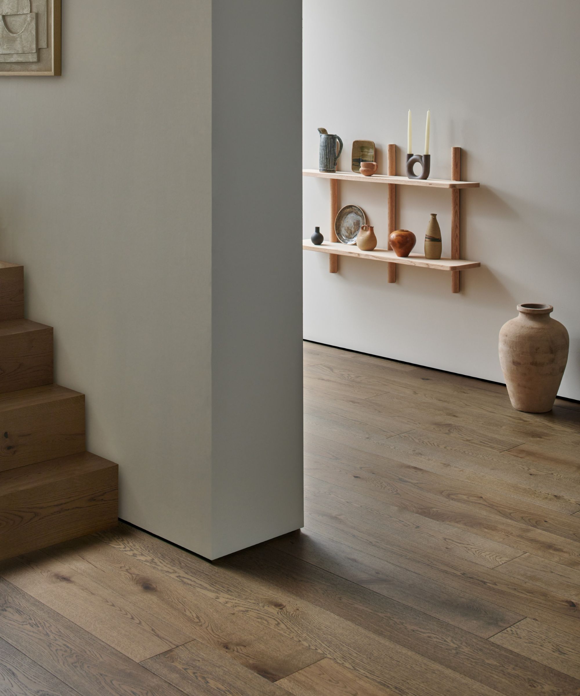 A quiet interior corner with warm wooden floorboards and a simple staircase. Pale walls hold two narrow wooden shelves displaying handmade pottery and candles. A large ceramic vase stands on the floor, creating a calm, uncluttered, gallery-like atmosphere.