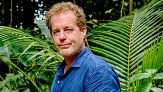 Rick (Adam James) stands in front of some palm fronts, wearing an ocean-blue cotton shirt