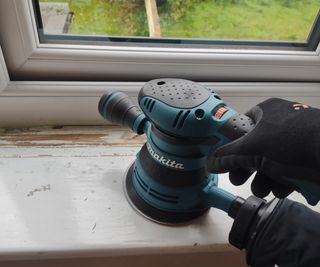 A random orbital sander being used on a windowsill removing a thick layer of white paint