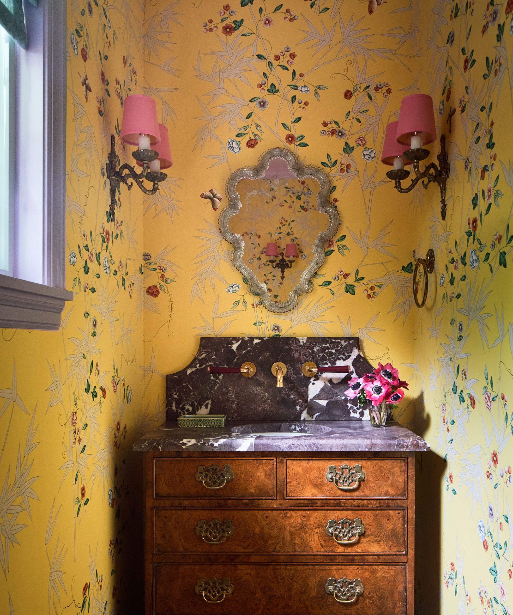 a yellow floral wallpapered powder room with antique wooden vanity with dark marble stone counters and shapely vintage mirror and sconces