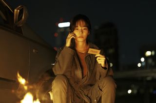 A woman holds a business card and talks on the phone while sitting by a fire and her car, outdoors at night, in 'Squid Game' season 2.