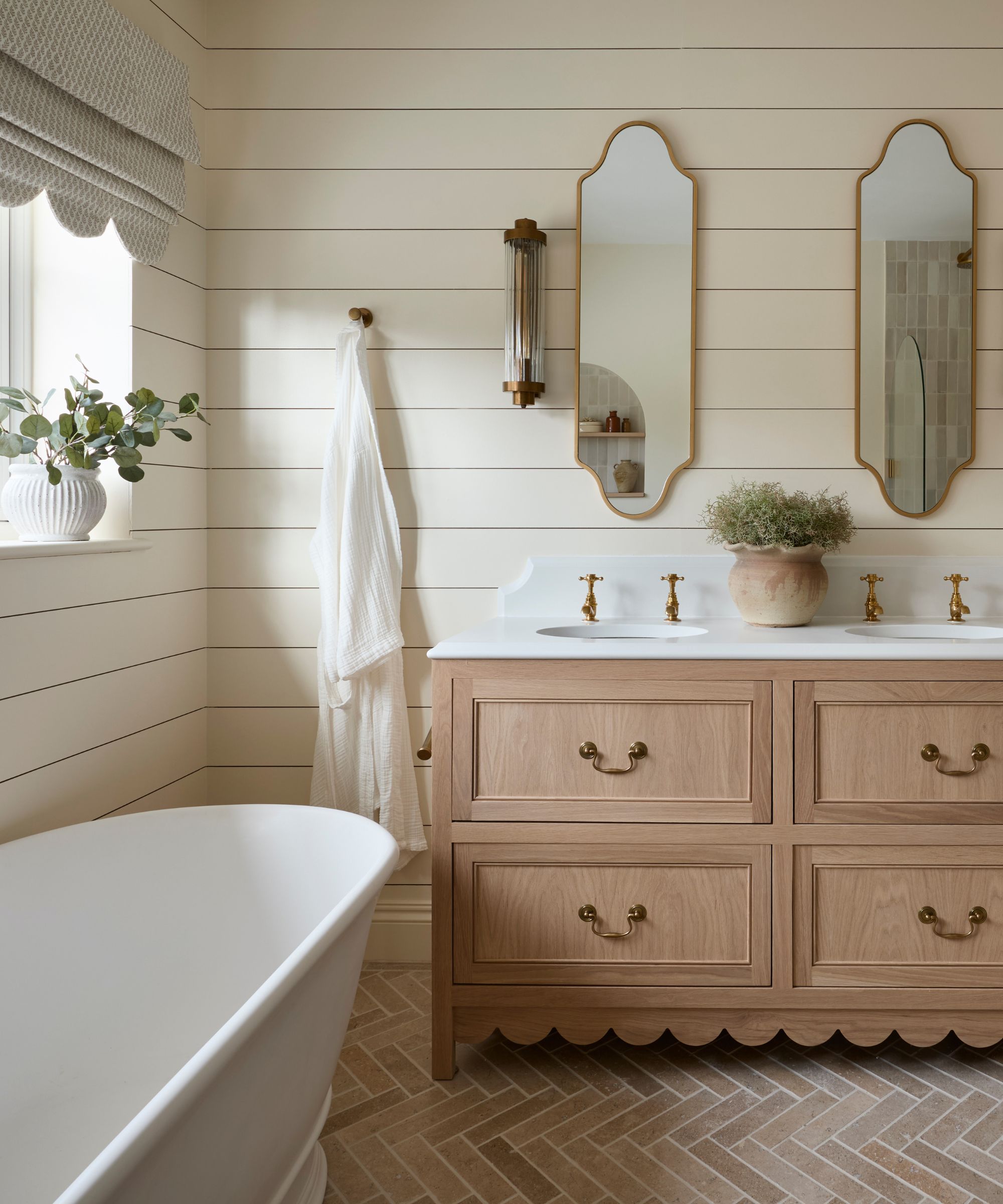 cottage country style bathroom with panelled walls, terracotta tiled floors, a wooden vanity unit with double sinks and a scalloped bottom trim with two vintage style mirrors and sconces
