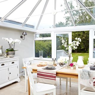 a wooden dining table with white wooden chairs inside a large airy conservatory with white walls and glass ceiling