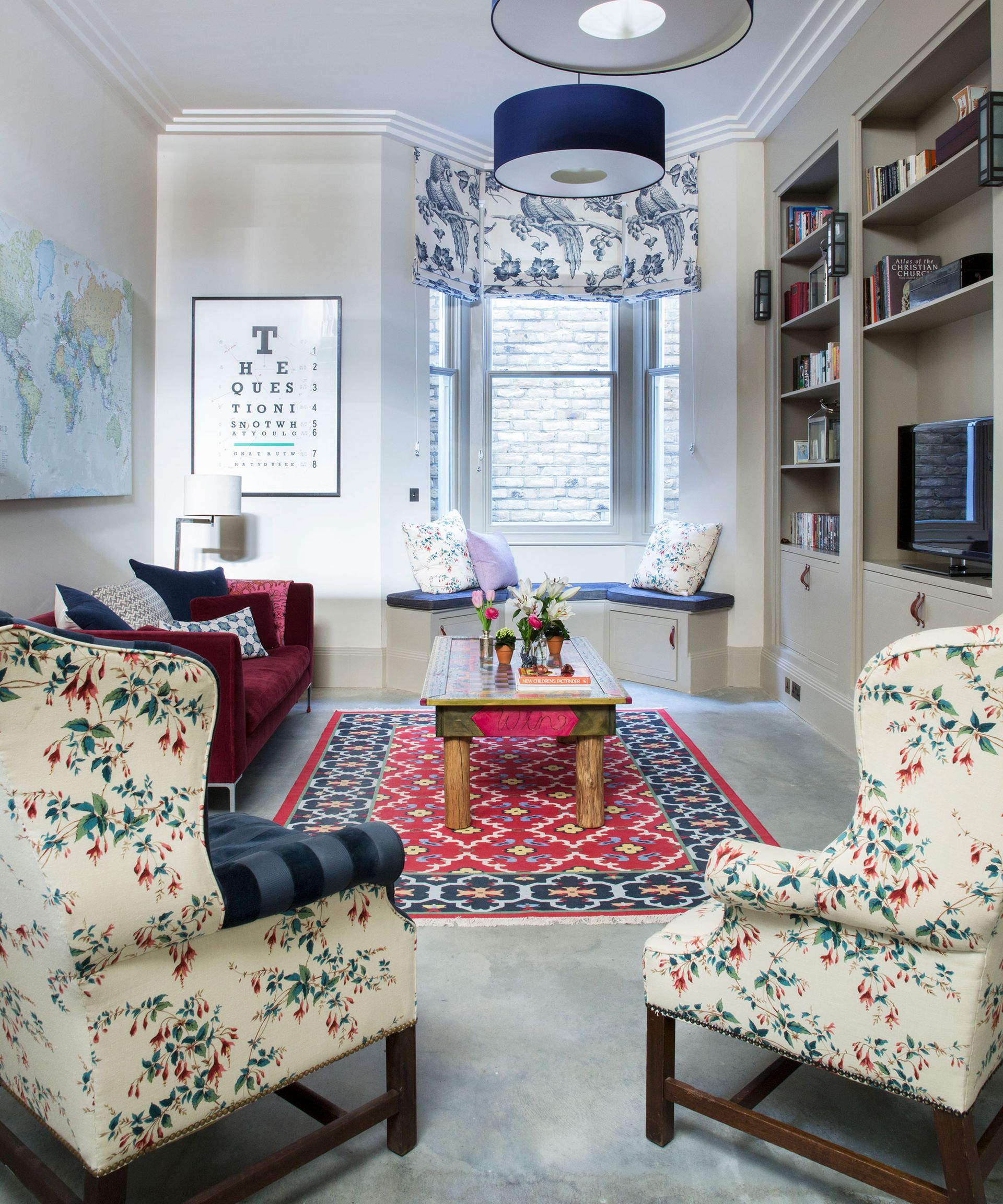 A long white living room with grey carpet, a brightly coloured rug and floral armchairs.