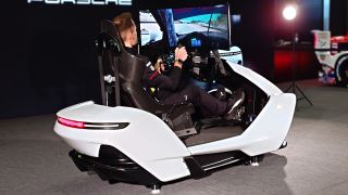 A photo of the ImSim Porsche Raceborn racing simulator in white showing a man driving in front of a black backdrop with the Porsche logo.