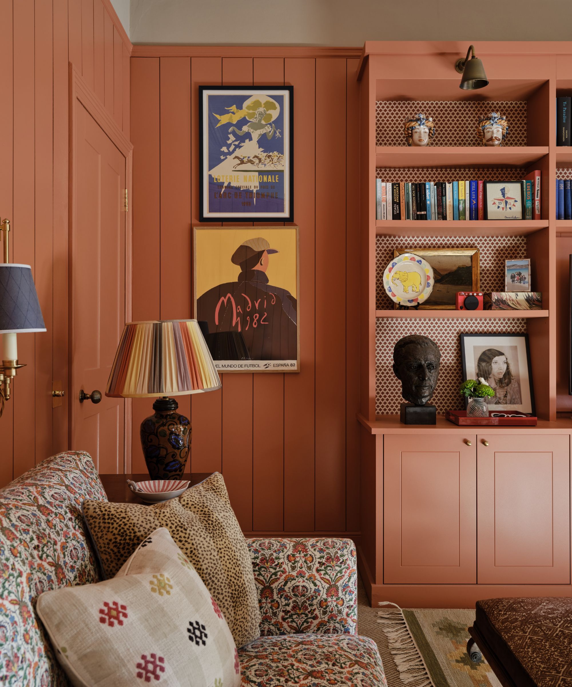 A cozy living room color-drenched in coral-red, with wall paneling and a built-in shelving unit. Eclectic decor on the shelves and two graphic, colorful artworks on the wall. A floral sofa with patterned cushions and a striped lampshade.