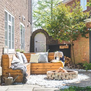 Wooden cornered seating around brick fire pit on gravel area in garden