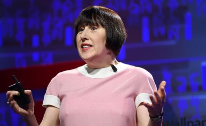 Alice Rawsthorn on stage wearing a pink top. Alice has short black hair and is using her arms when giving a speech. 