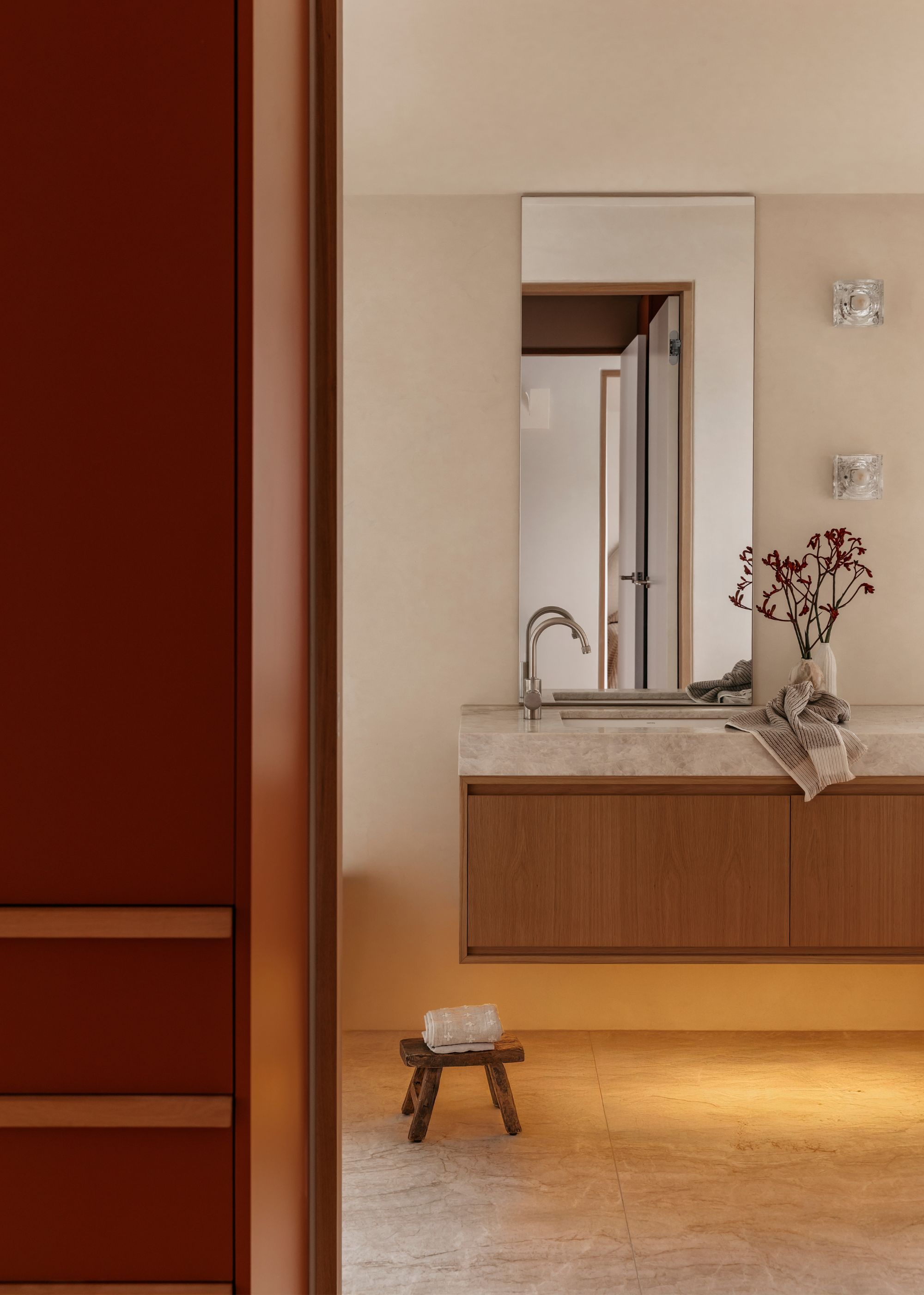 A bathroom with a long mirror, a floating basin, a vase of flowers, a pair of glass sconces, and a mini stool with a towel