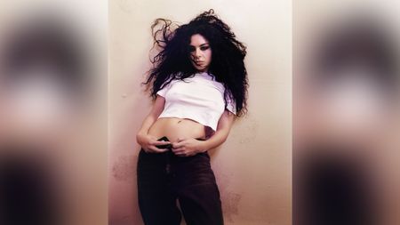 Charli xcx leans back against a pale wall wearing a white crop top and dark jeans, her dark curly hair caught mid-movement, her gaze directed downward; photographed by Harley Weir, 2024.