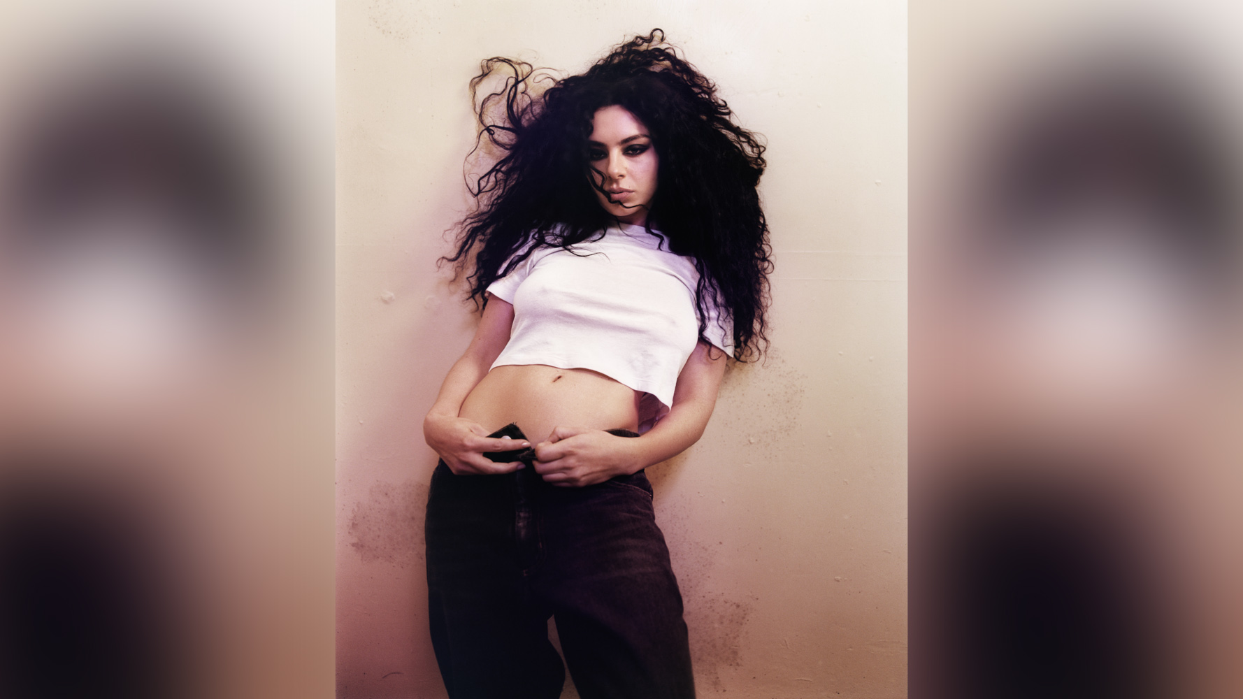Charli xcx leans back against a pale wall wearing a white crop top and dark jeans, her dark curly hair caught mid-movement, her gaze directed downward; photographed by Harley Weir, 2024.