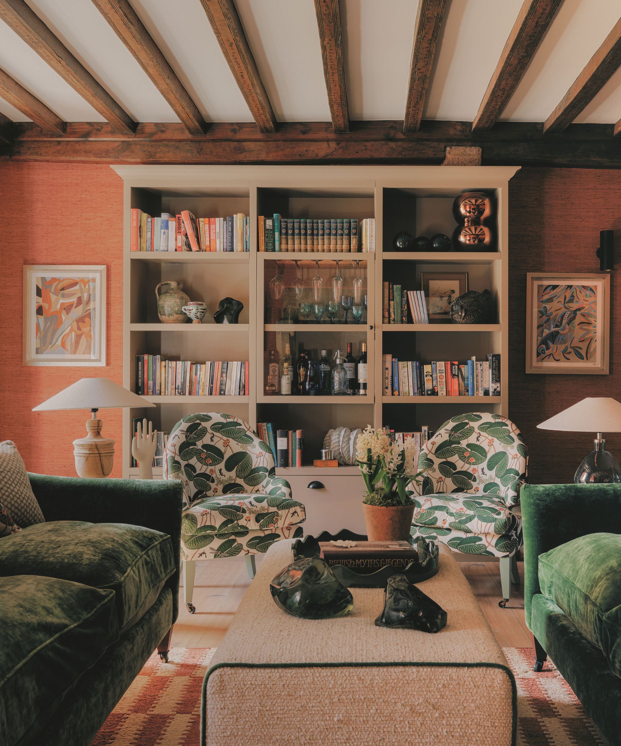 Living room view showing green velvet sofas, patterned armchairs, a central ottoman coffee table, exposed ceiling beams, and filled bookshelves.