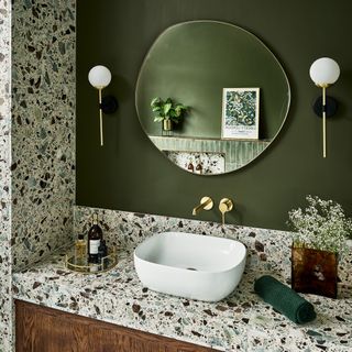 a green bathroom with a vanity area with a single white basin, terrazzo wall and countertop, wooden floating vanity unit, globe wall lights and round wall mirror