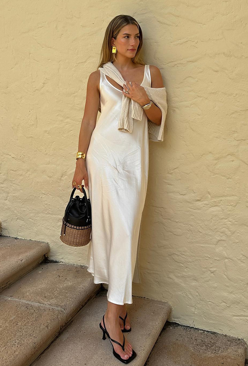 A woman's white dress outfit with slip styled with cardigan, raffia bag, and black heeled thong sandals.