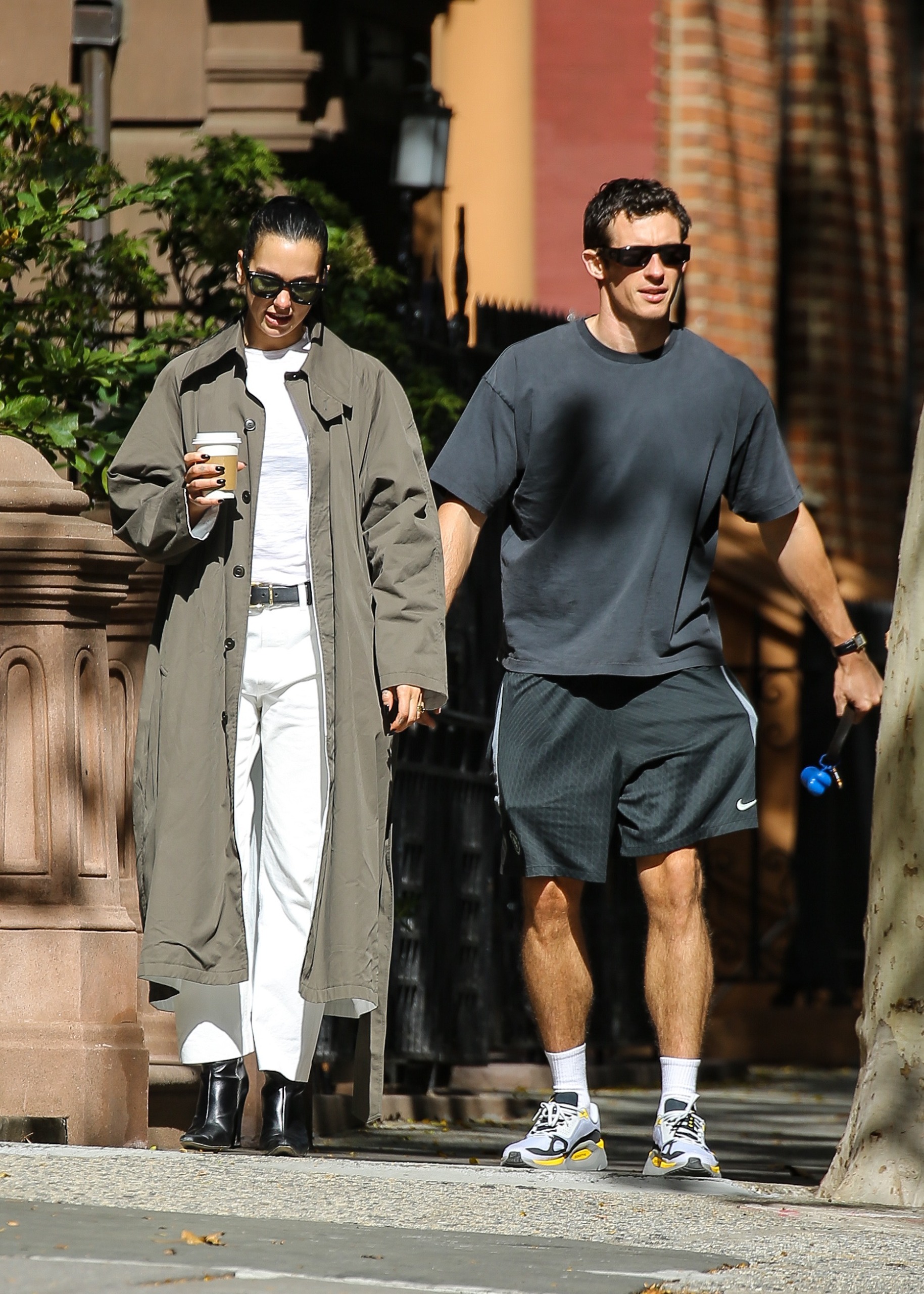 Dua Lipa wears white pants with an anti trend trench coat and black boots while walking with husband Callum Turner