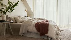 White, beige, and brown bedding on a bed against white walls and white sheer curtains; a nightstand to the left of the bed, topped with a potted plant. 