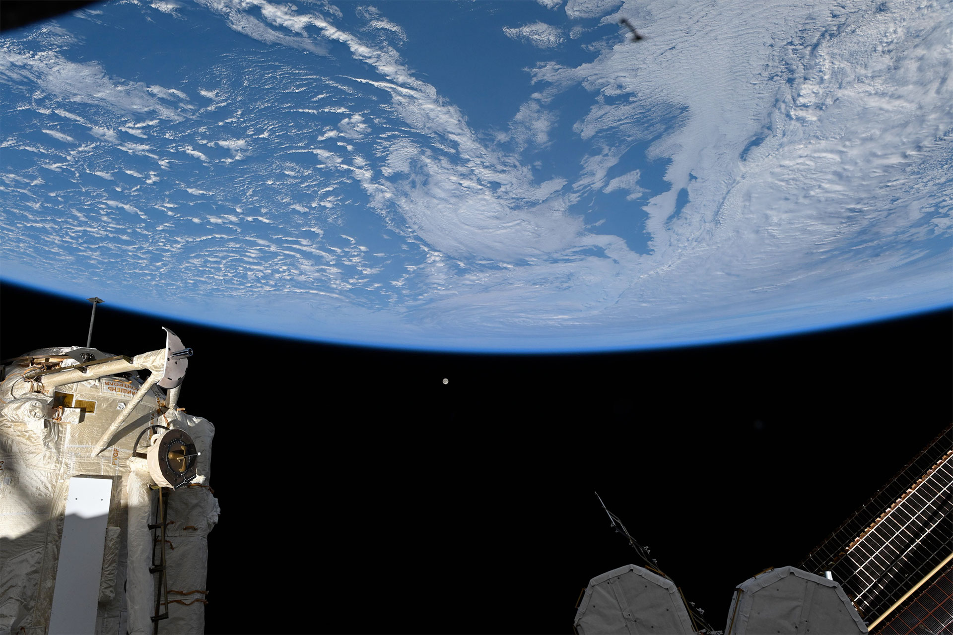 a full moon looks tiny set against the backdrop of the blackness of space with Earth's horizon and a space station in the foreground..