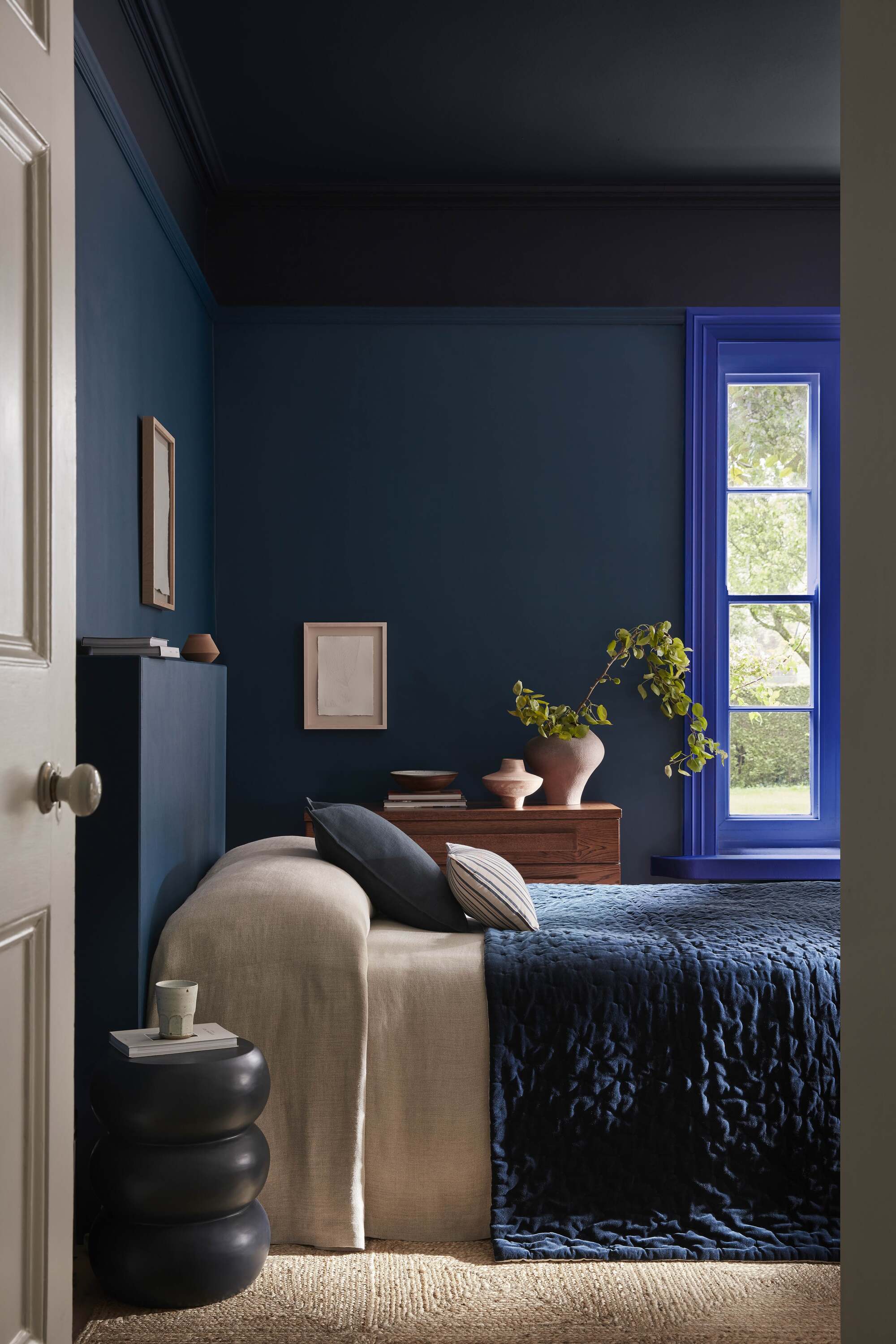 modern bedroom color drenched in navy blue with cobalt blue window frame, blue quilt, with beige sheets, a jute rug, and a cabinet with a vase with flowers on the opposite wall