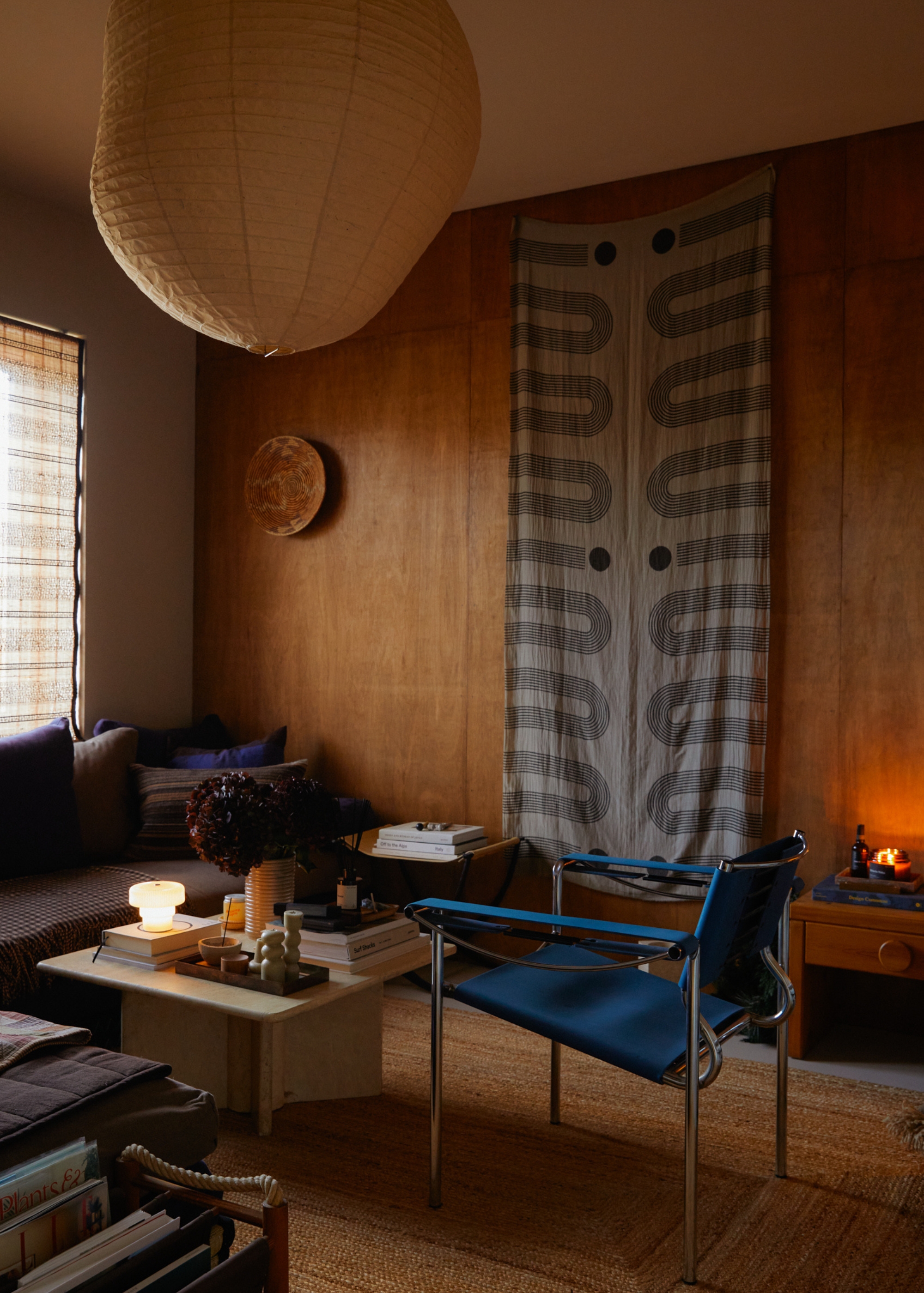 A living room with a paper pendant lamp, a blue chair with metal accents, a travertine coffee table, a patterned fabric wall hanging, a vase of flowers, a mini lamp, and a candle