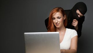 Red-headed white woman types on laptop while menacing figure wearing balaclava looks over her shoulder.