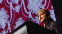 A low-angle shot shows a person wearing a patterned head covering and a denim jacket speaking from behind a dark podium, with a large screen displaying a red and white floral pattern blurred in the background.