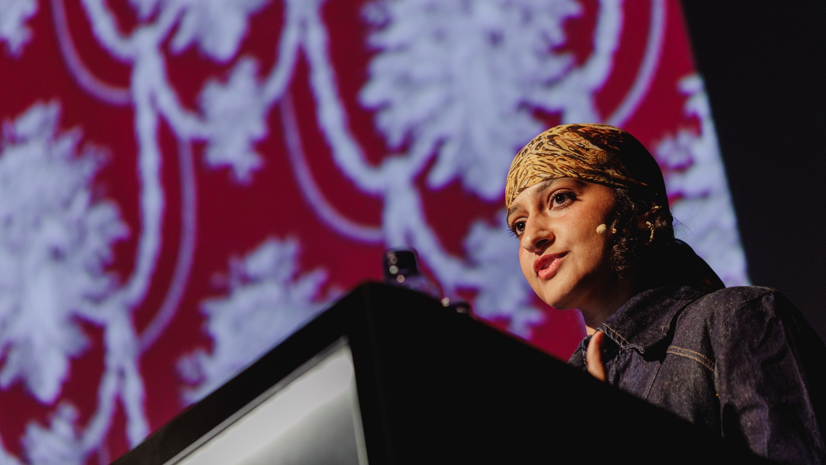 A low-angle shot shows a person wearing a patterned head covering and a denim jacket speaking from behind a dark podium, with a large screen displaying a red and white floral pattern blurred in the background.