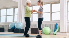 Personal trainer holds hands of older white-haired woman balancing on wobble board