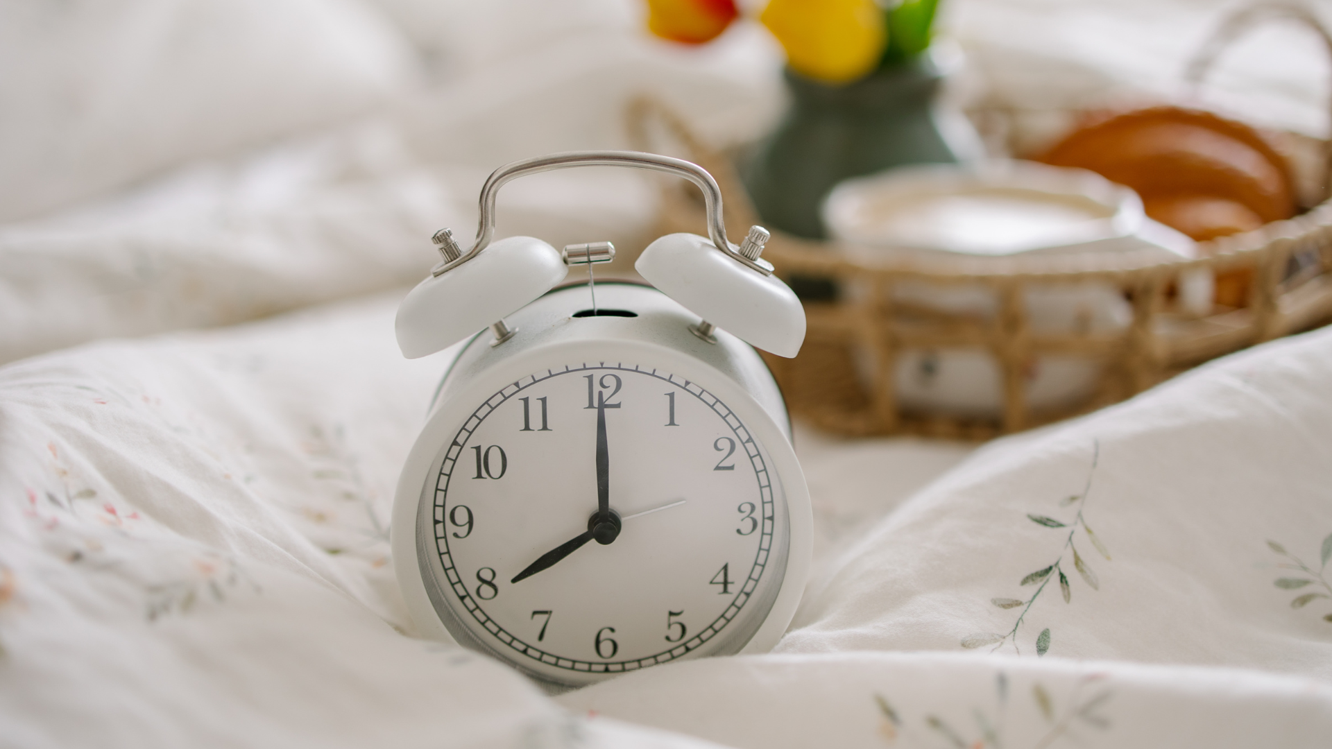 A white alarm clock rests on a bed in a sunny room.