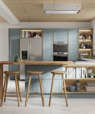 A kitchen with a long wooden island, three wooden bar stools, a shelf of books, and light blue cabinets on it and behind it as well as a silver fridge and two ovens stacked vertically in the cabinet