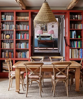Country modern dining room with wooden beams and white ceiling, red fitted storage shelves with books and ornaments, light wood dining table with matching dining chair, textured, woven pendant over dining table