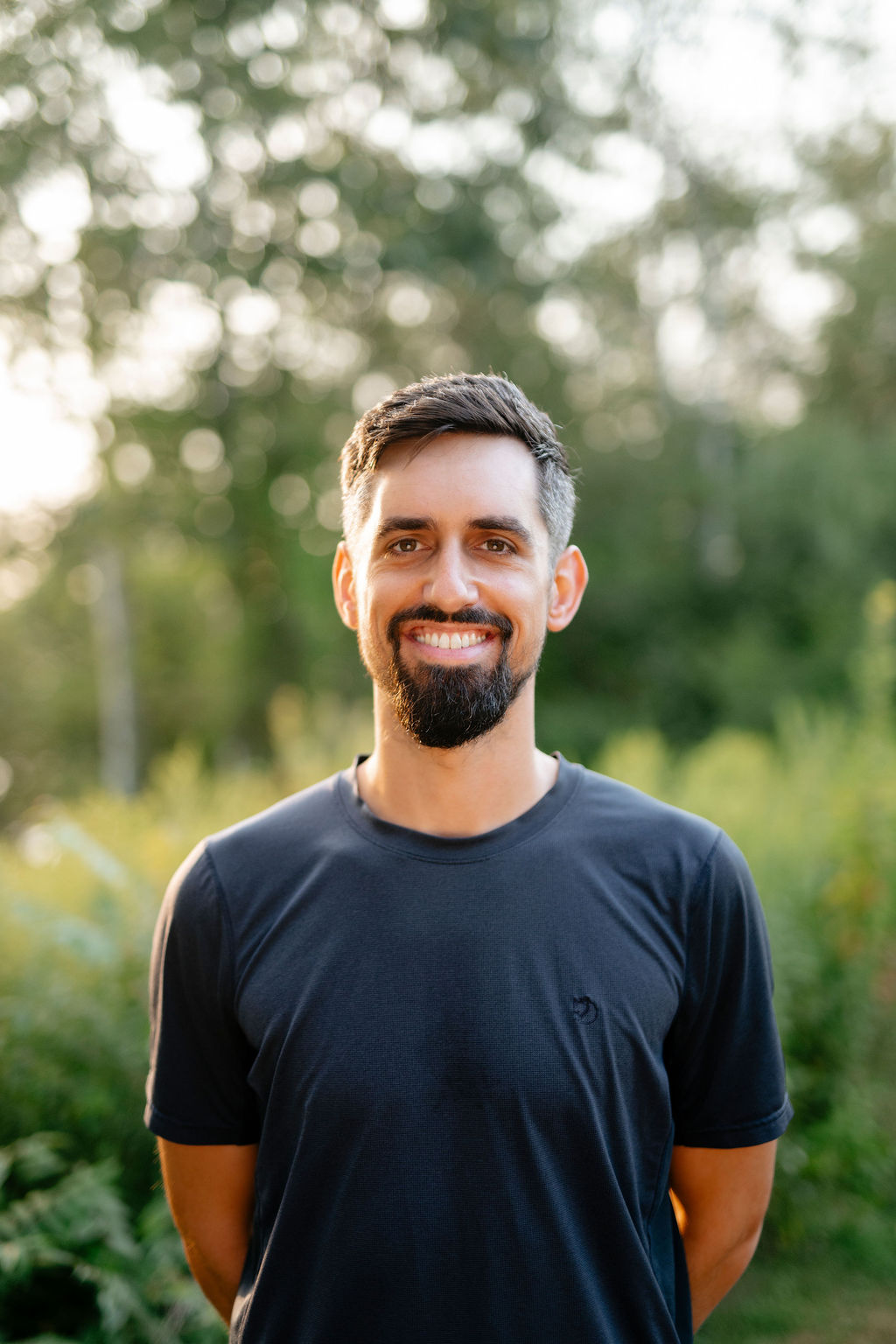 man facing the camera smiling in a black tshirt with trees blurred in the background