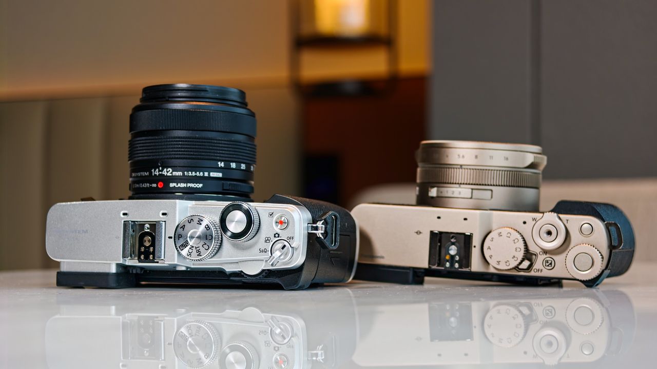 OM System PEN vs Panasonic Lumix L10: two micro four thirds cameras side by side on a white marble surface with lamp light in the background
