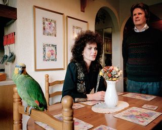 A kitchen with 2 framed wall art on a white wall. A parrot sittin on the head of a chair in the dining area. A woman sitting at the dining table on a chair with a man standing closeby. Flowes in a white vase in the middle of the brown dining table with table mats laid