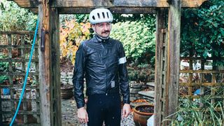 A white man wears a black rapha shakedry waterproof cycling jacket under a trellis