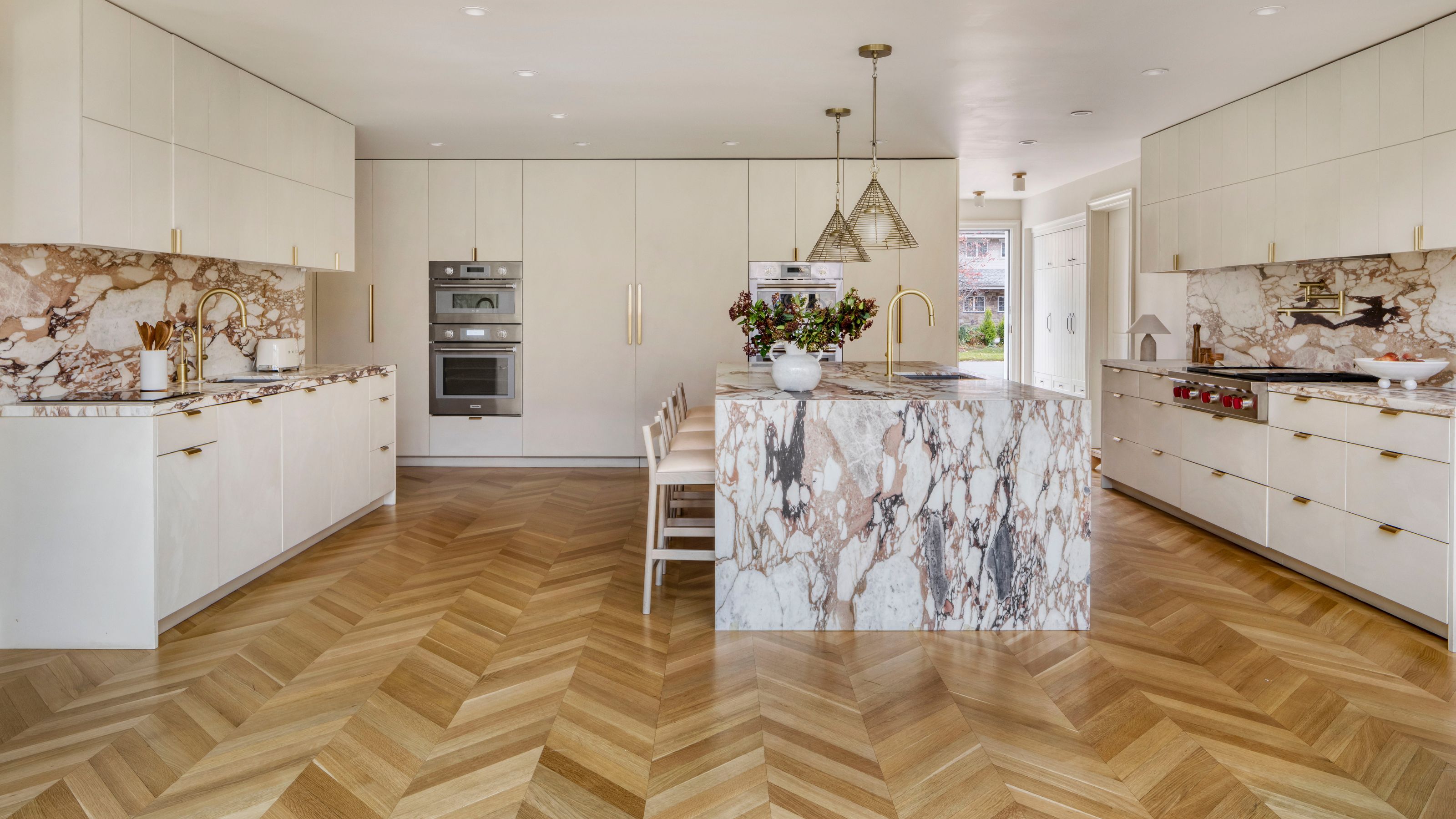 A bright and airy kitchen with a large marbled island in the middle as well as white cabinets and gold pendant lights hanging. There is also a view of the integrated oven and microwave as well as other elements of the kitchen