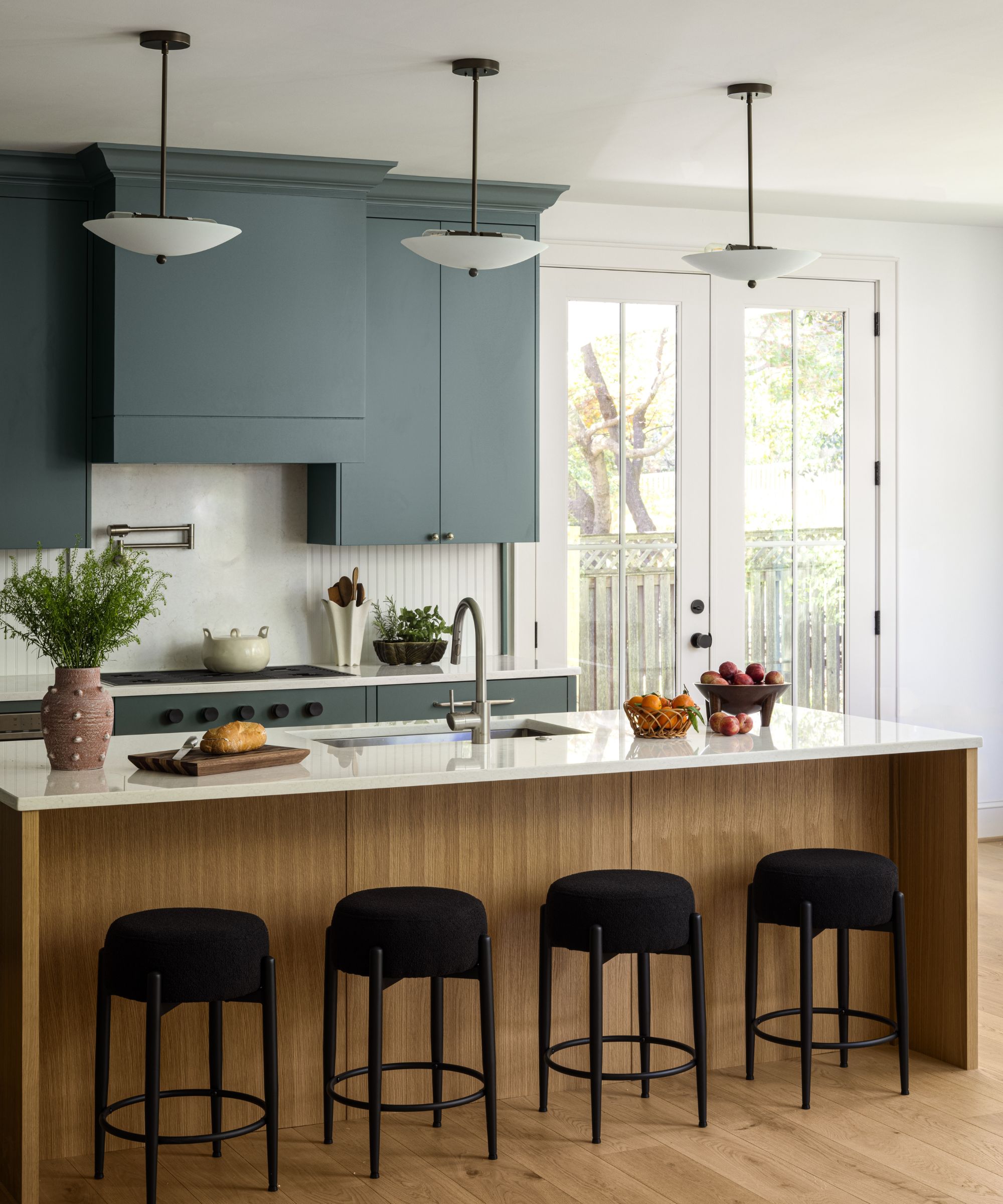 A modern kitchen with blue-gray cabinets, white walls, a wooden kitchen island with black bar stools, wooden flooring, and French doors leading outside.