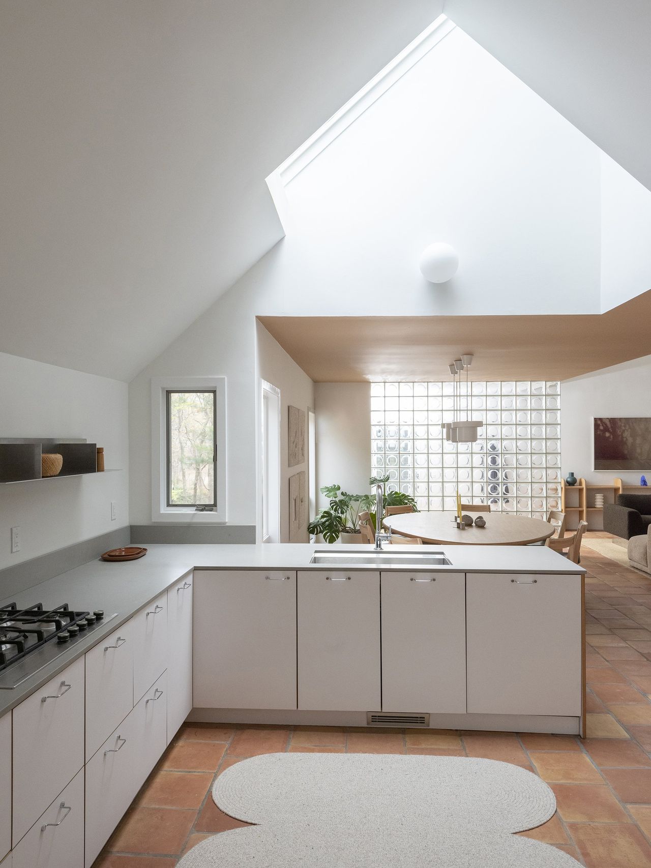 A modernly designed open-air kitchen area features predominantly white furniture, wooden floor decorated with a white rug, a round wooden table, and an accent glass wall, as well as a triangularly cut ceiling.