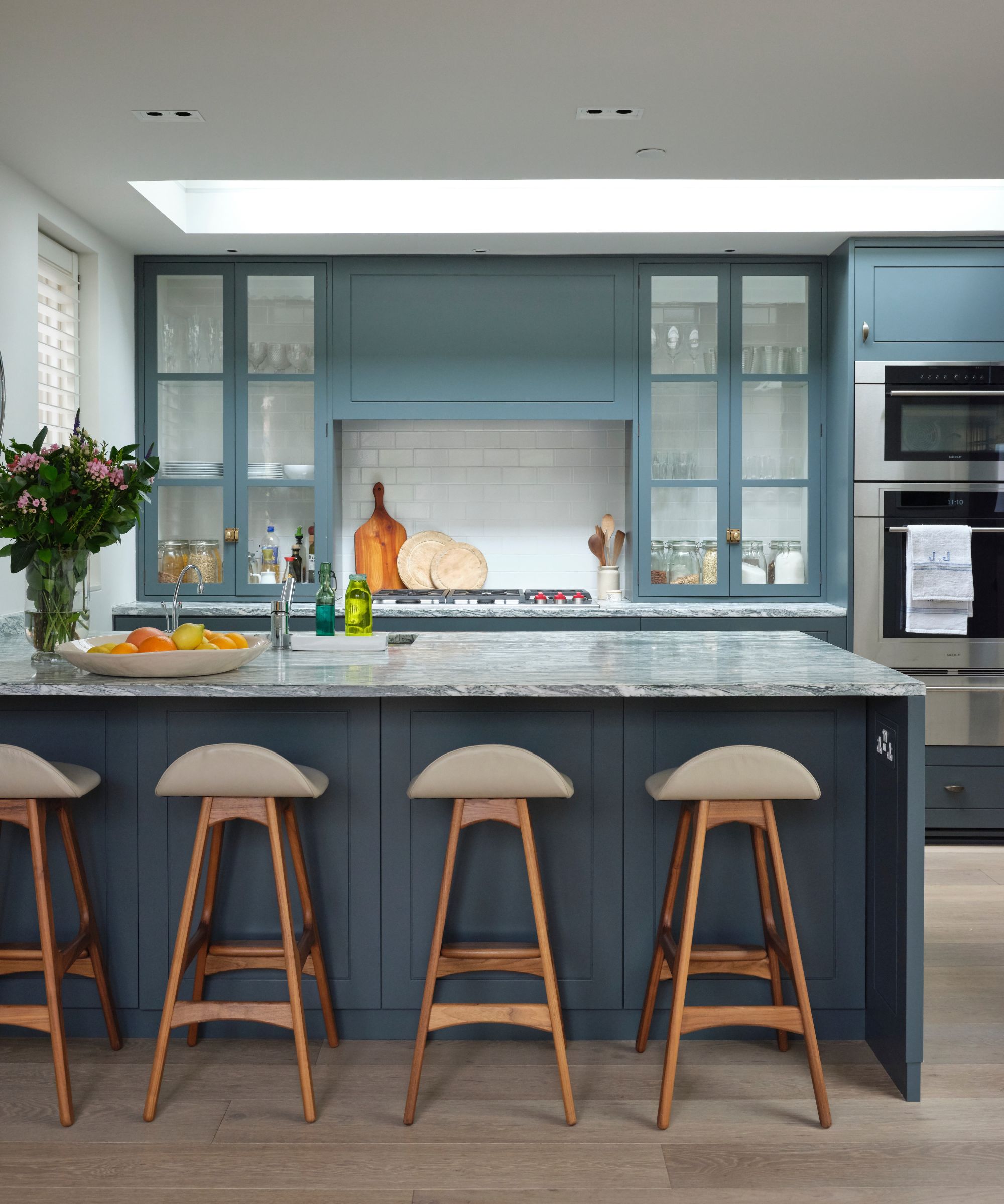 A blue kitchen with a blue kitchen island with marble countertop, wooden bar stools, glass fronted cabinets and white tiled backsplash.