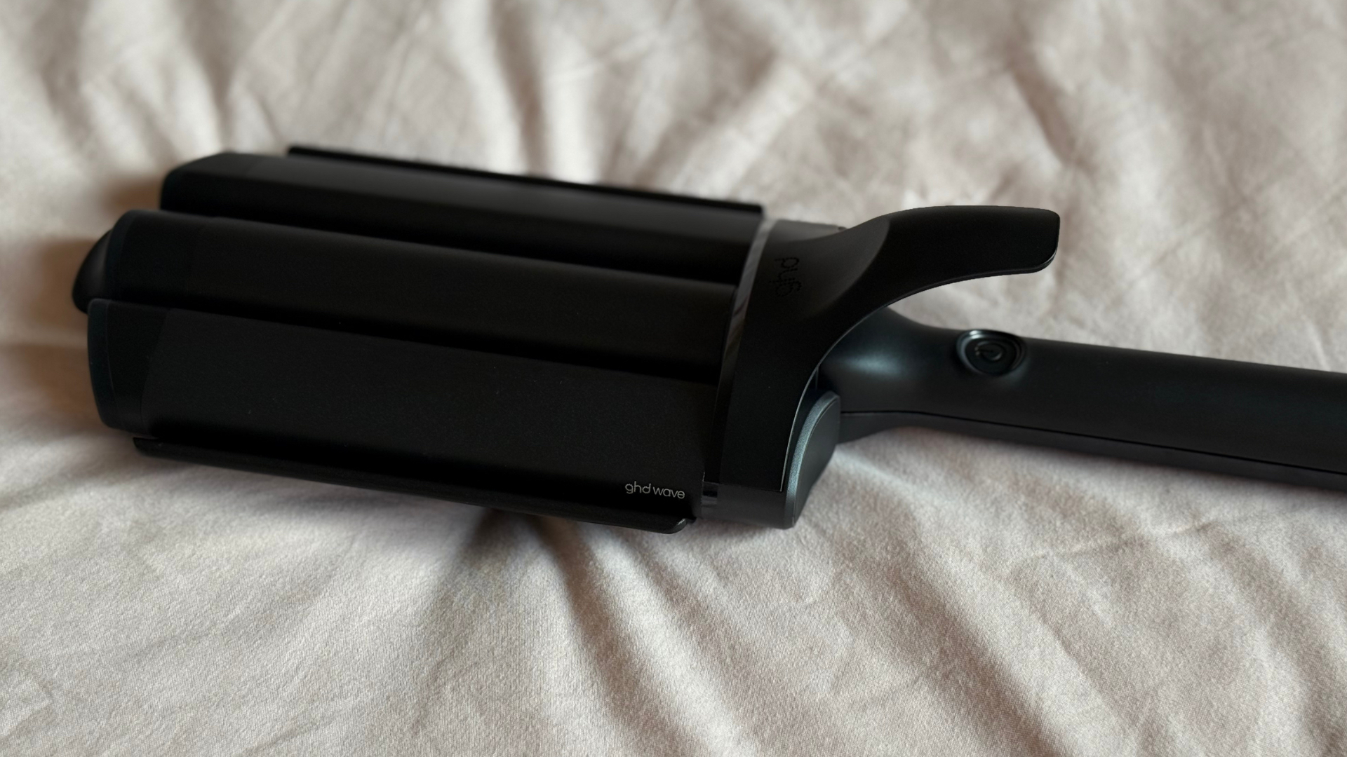 A side view of the ghd Wave Triple Barrel Waver Tool laying on a dusky pink fabric background