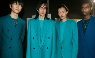 Four models wearing looks from Paul Smith's collection. One model is wearing a green top, chain with pendants and teal suit jacket. Two models are wearing sea blue tops and suit jackets. And the fourth model is wearing a dark blue top, chain with pendant and dark blue suit jacket