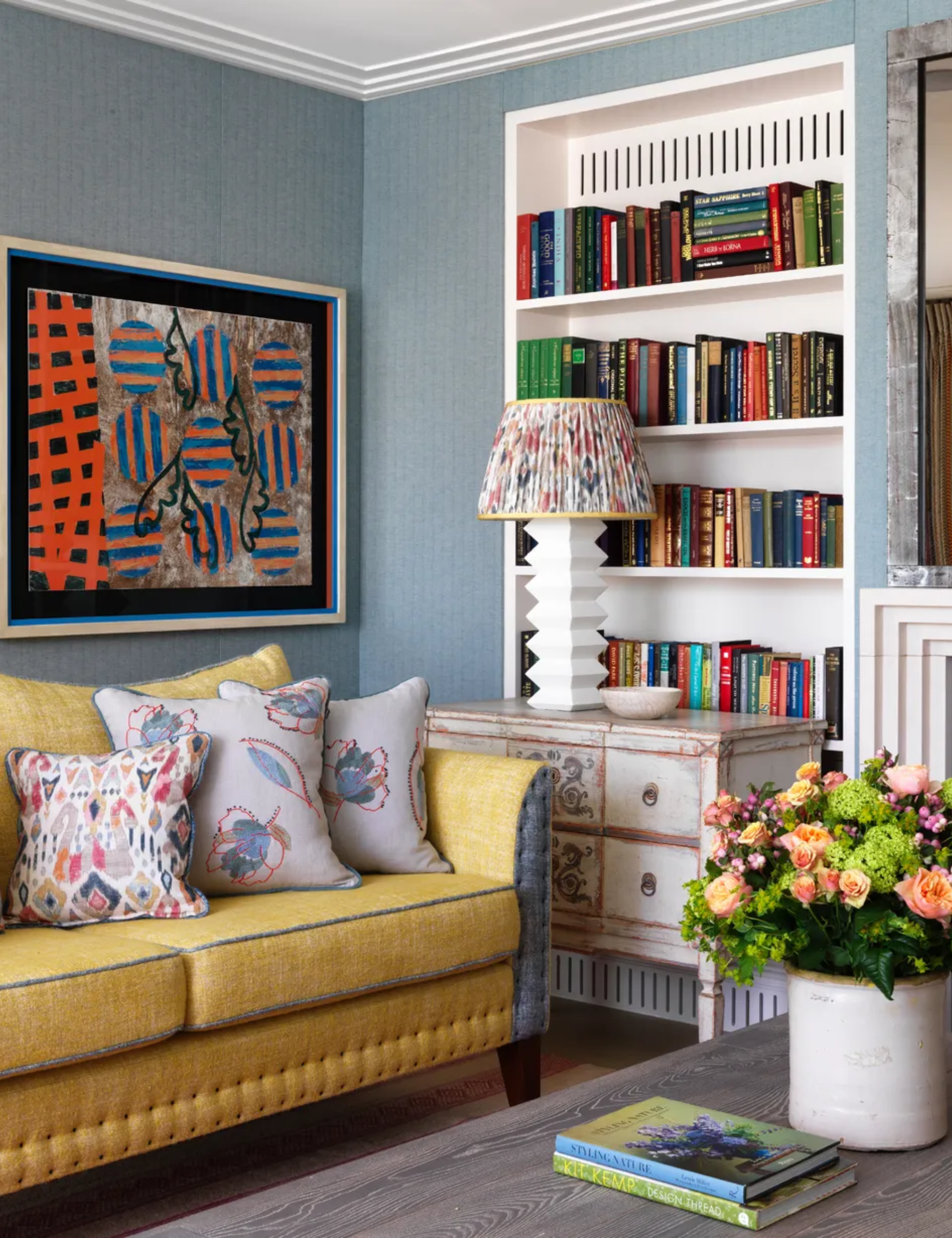 blue living room with yellow couch, patterned couch pillows, ornate sideboard, bookcase, modern artwork, flowers, coffee table