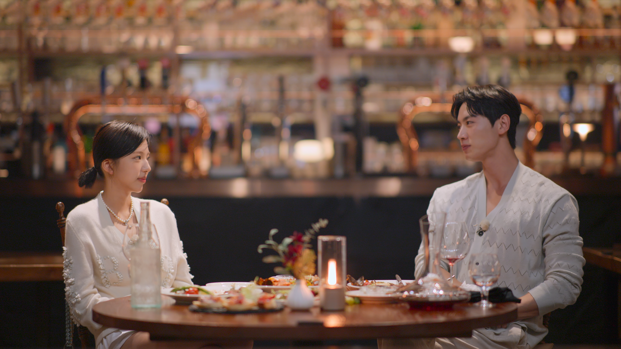 A woman (Kim Go-eun) and a man (Jo I-geon), both wear white as they have a meal together. The fully-stocked bar behind them is blurred, in a still from 'Single's Inferno 5.'