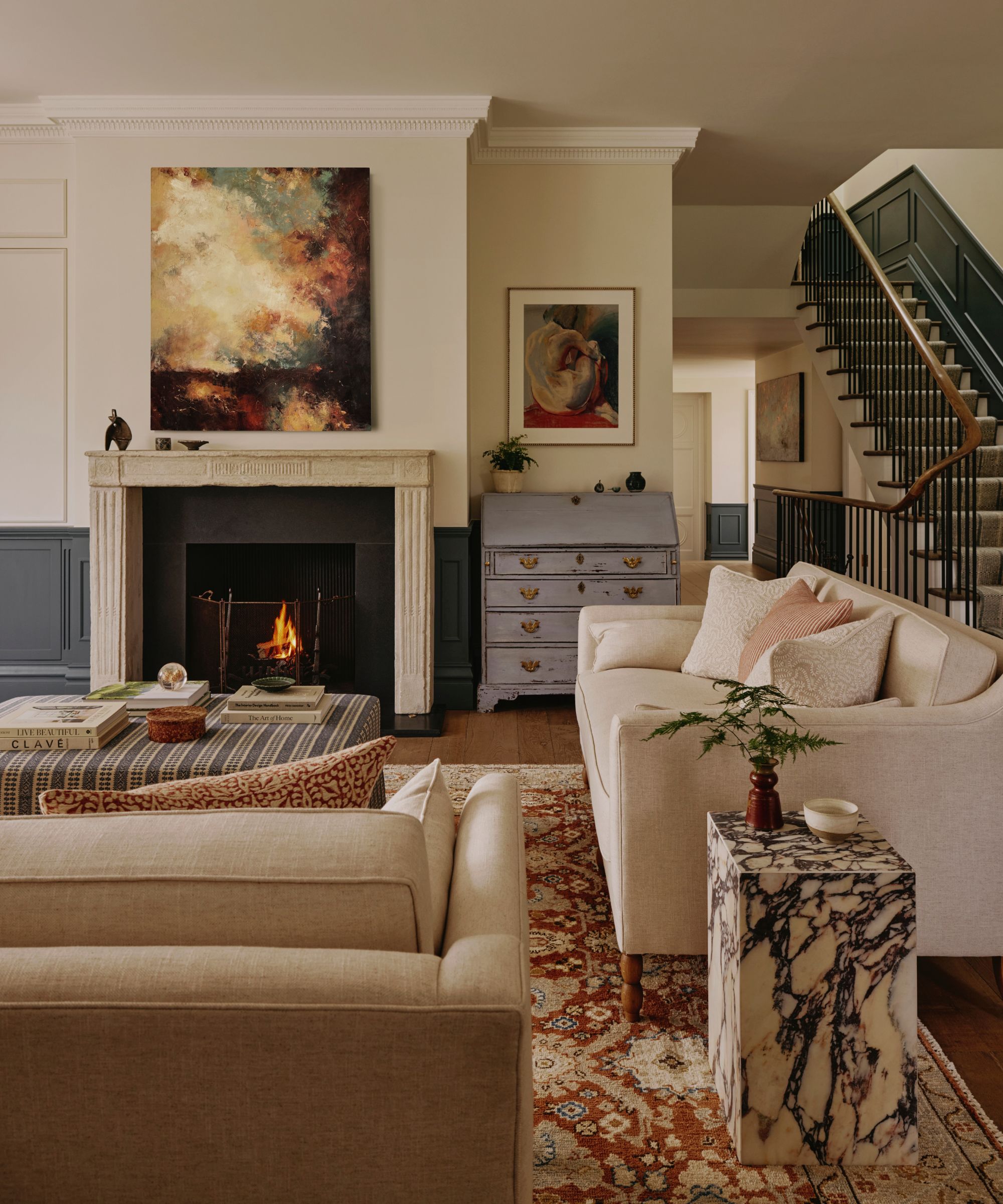 neutral living room with two tone paneling, original wooden fireplace, statement abstract art, two neutral couches and a striped ottoman