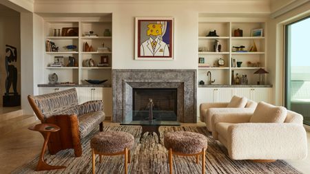 Modern transitional living rooms with cream walls, alcoves with bookshelves filled with decor and trinkets, a large fireplace with a Pop Art print above, two boucle armchairs, and a vintage sofa.