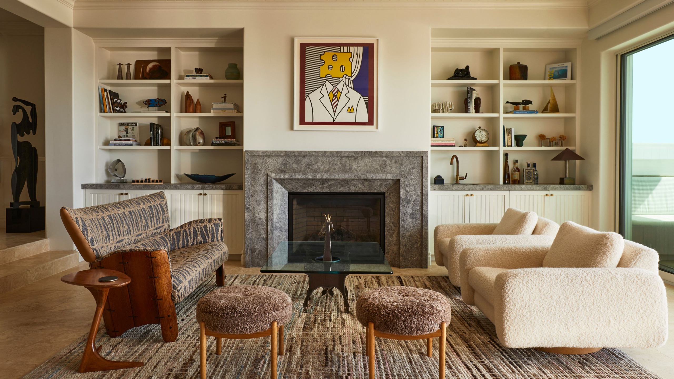 Modern transitional living rooms with cream walls, alcoves with bookshelves filled with decor and trinkets, a large fireplace with a Pop Art print above, two boucle armchairs, and a vintage sofa.